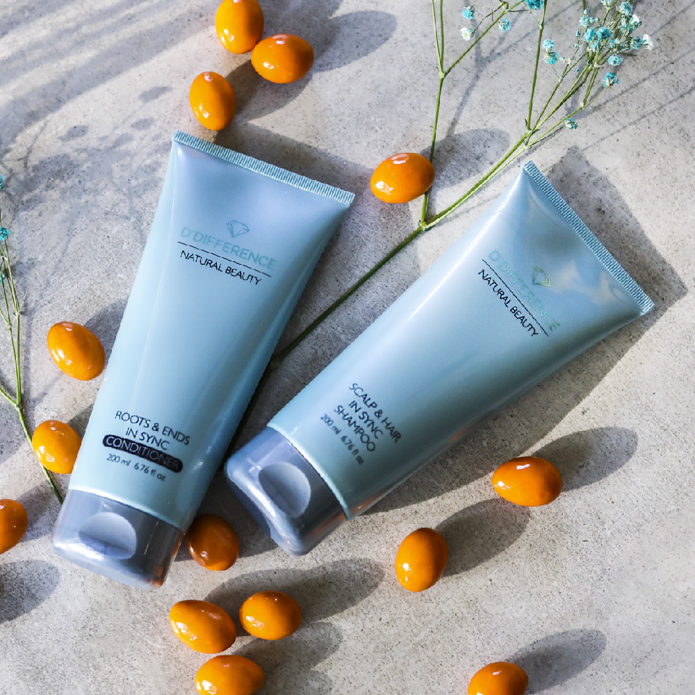 Blue tubes of conditioner and shampoo from D'DIFFERENCE on concrete with orange pebbles and some green plants, all in sync